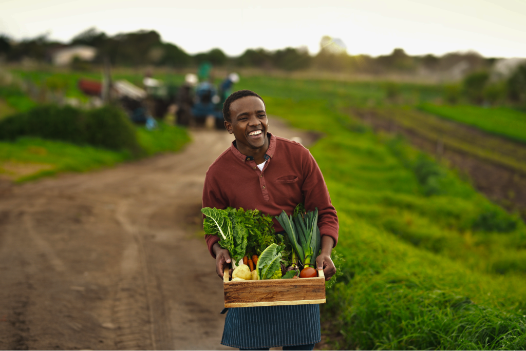 A farmer smiling and carrying a crate of fresh harvest vegetables